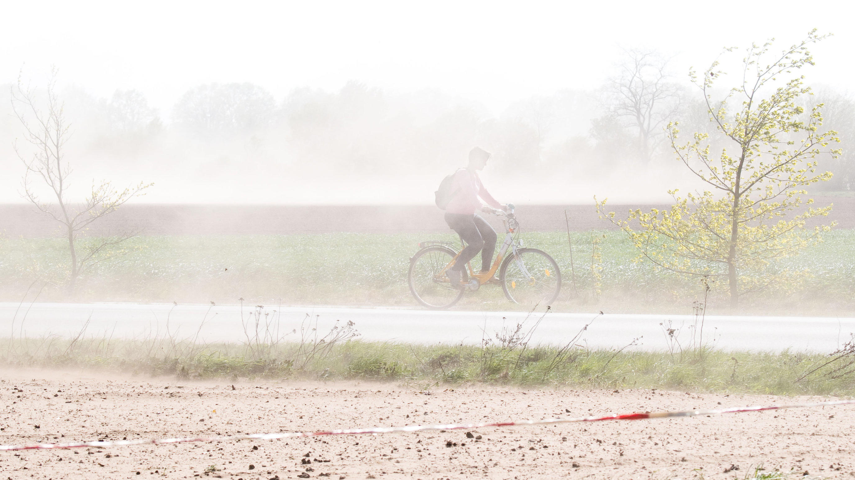 „Sandsturm“: Kräftiger Wind fegt in Norddeutschland über ausgetrocknete Böden 23.04.2019, Niedersachsen, Burgdorf: Eine Fahrradfahrerin fährt mit ihrem Rad über einen Fahrradweg, als starker Wind viel Sand von trockenen Feldern aufwirbelt. Foto: Julian Stratenschulte/dpa +++ dpa-Bildfunk +++
