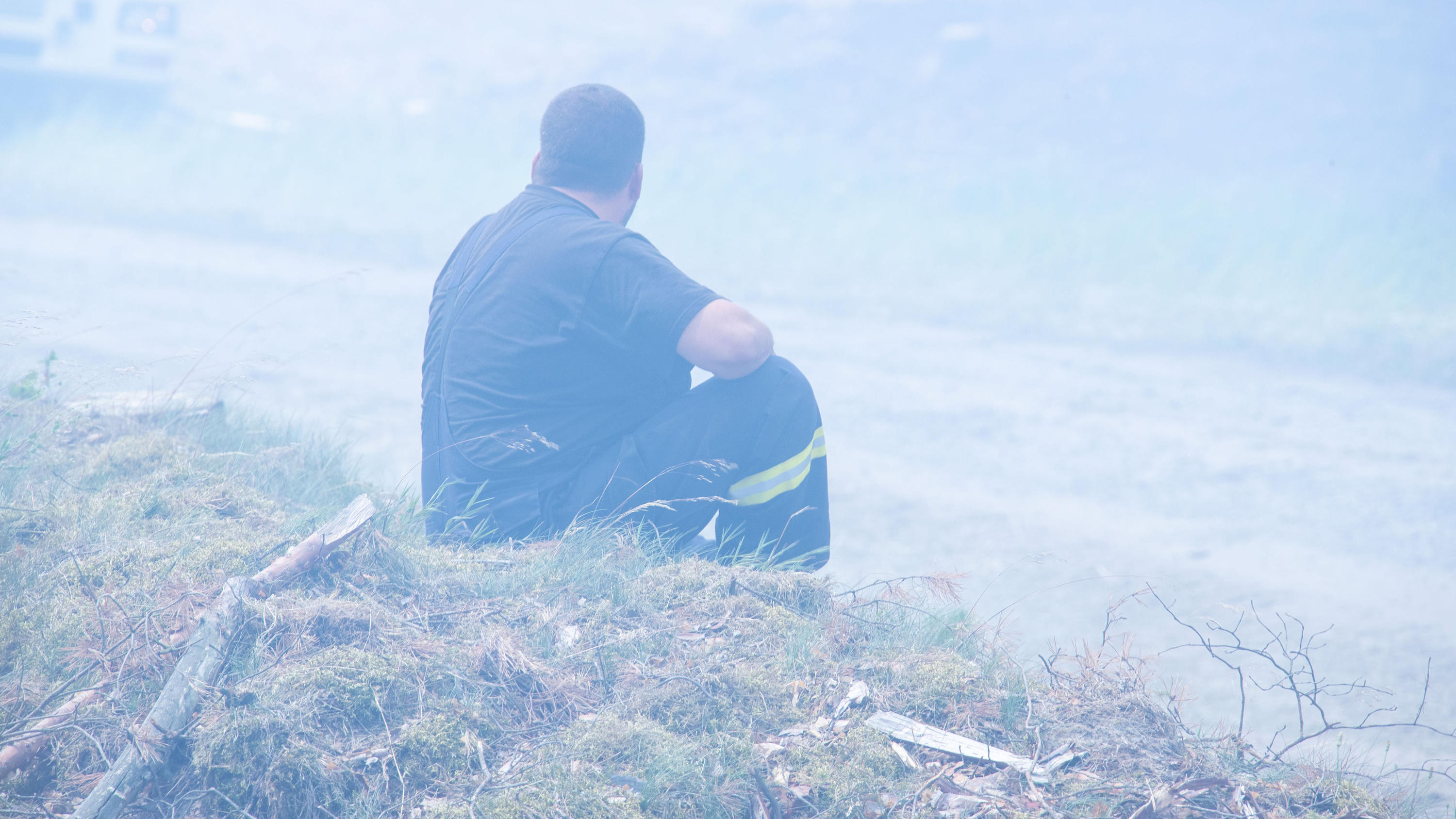 Vor einem Jahr wütete in Lübtheen das Feuer – jetzt herrscht wieder höchste Waldbrandgefahr 01.07.2019, Mecklenburg-Vorpommern, Alt Jabel: Ein erschöpfter Feuerwehrmann sitzt in einer Rauchwolke im brennenden Wald in der Nähe der kleinen Ortschaft Alt Kabel am Rande des ehemaligen Truppenübungsplatzes bei einem großflächigen Waldbrand in de