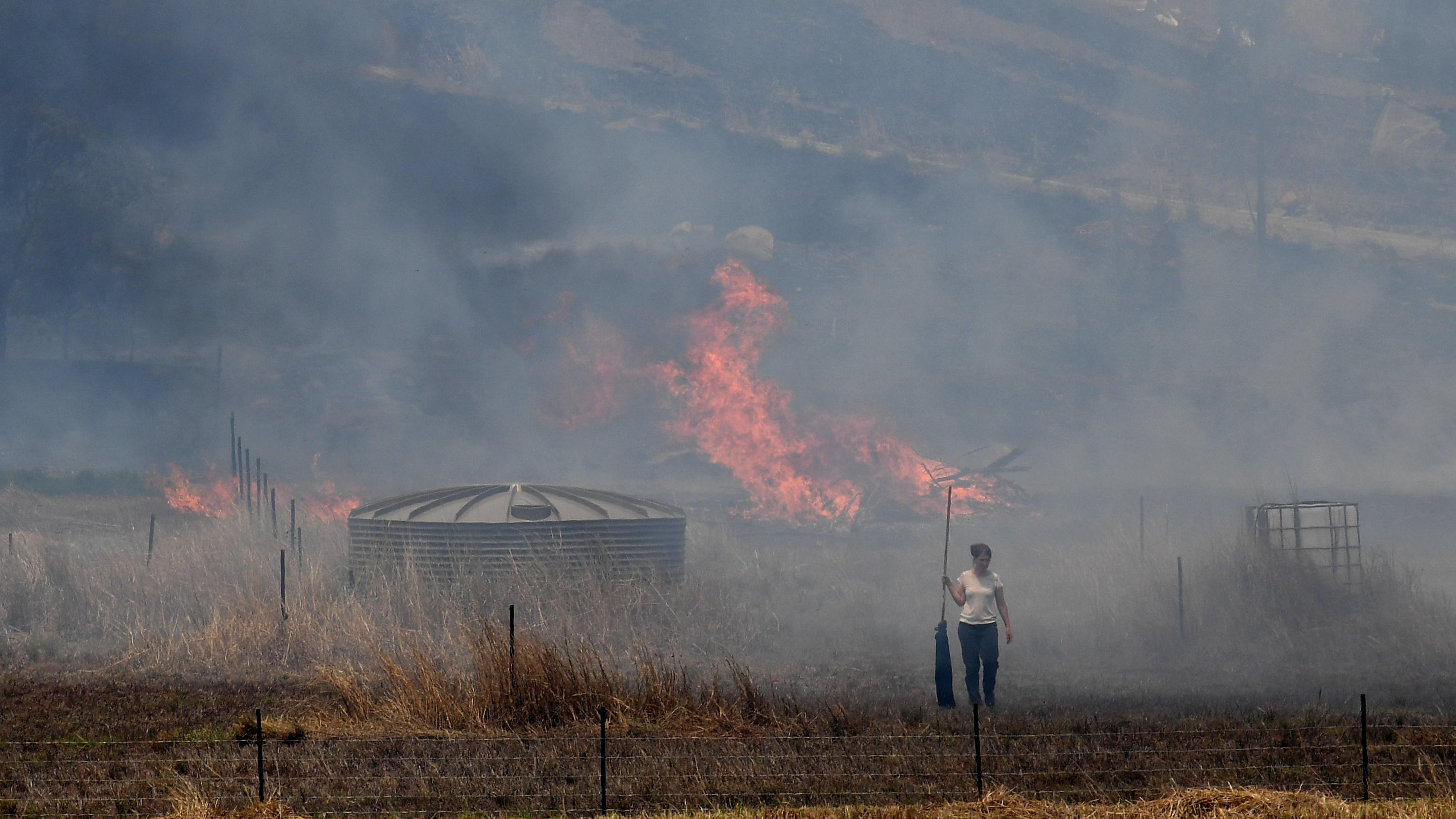 Der Faktencheck: Kriegen wir noch die Klima-Kurve? 13.11.2019, Australien, Taree: Eine Frau geht über ein Feld, während im Hintergrund Flammen eines Buschfeuers lodern. Die verheerenden Buschbrände im Osten Australiens haben nach Schätzungen der Feuerwehren bereits über 1,1 Millionen Hektar Land in