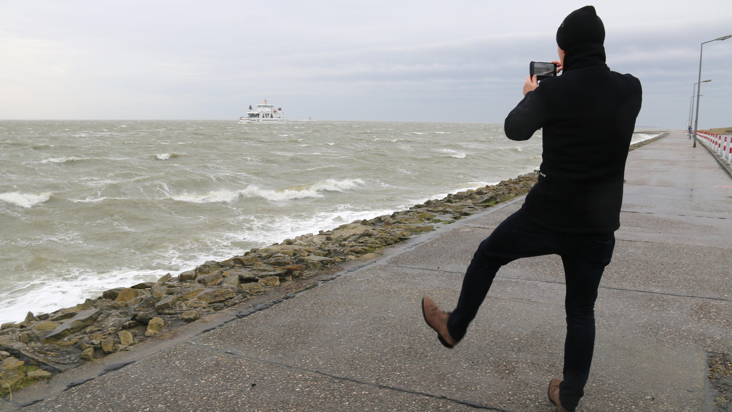Live-Ticker zum Orkan Sabine: Frau aus dem Main gerettet - Vier Rettungskräfte in NRW verletzt 09.02.2020, Niedersachsen, Norderney: Ein Mann macht auf der Strandpromenade auf der Nordseeinsel Norderney ein Foto von der vorbeifahrenden Autofähre. Die ersten Ausläufer des Sturmtiefs