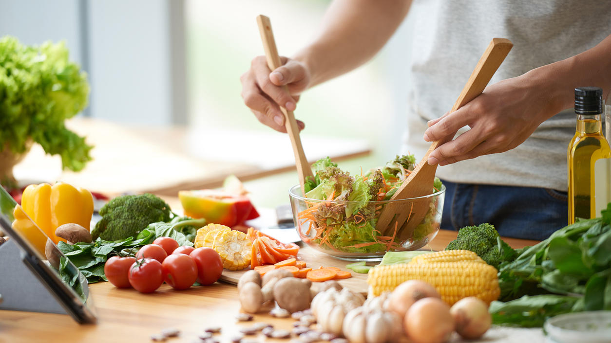 Hat die Maske wirklich unser Immunsystem geschwächt? Ein Mann mixt in einer Salatschüssel seinen Salat, um ihn herum liegen verschiedene Gemüsesorten.