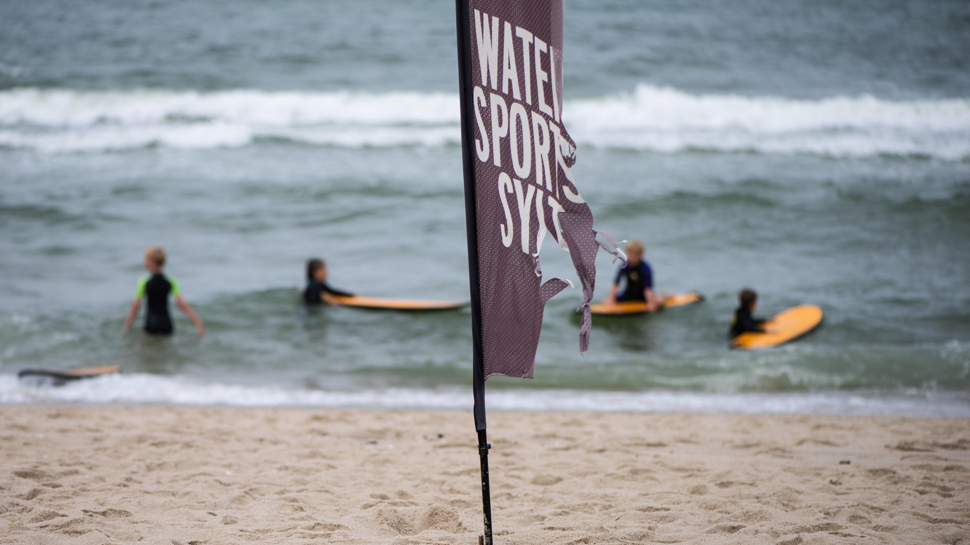 Wetterbericht für den 15.08.2020: Gewitter und Regenschauer kühlen die Nacht ab Junge Wassersportler am Strand 02.08.2020, Wenningstedt --- Wassersportler mit ihren Wellenreiter-Boards stehen in der Nordsee auf der Insel Sylt hinter einer Flagge Water Sports Sylt. Wenningstedt Schleswig-Holstein Deutschland *** Young water spor