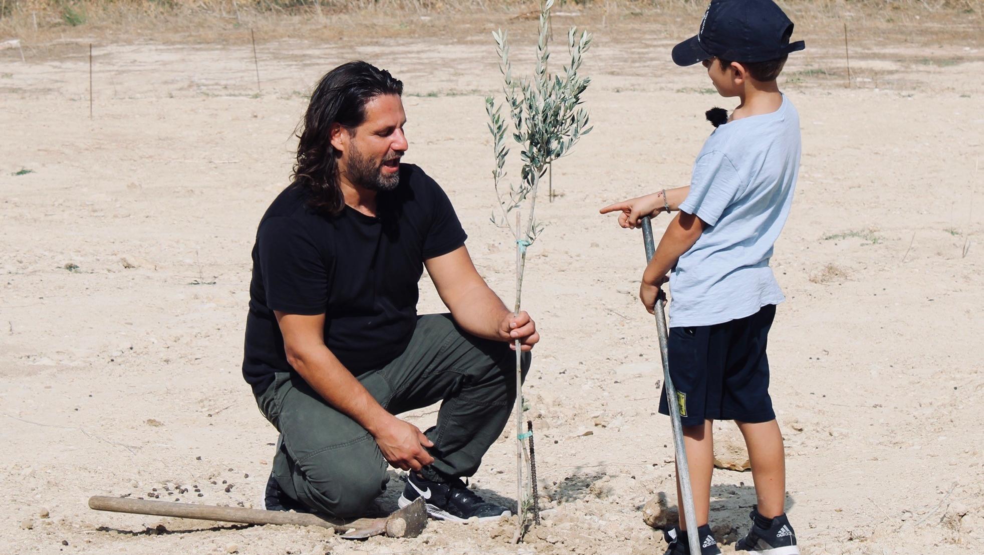 Baumpate werden und auf Kreta alte Olivenhaine schützen Adam Bousdoukos und kleiner Junge knien vor junger Pflanze