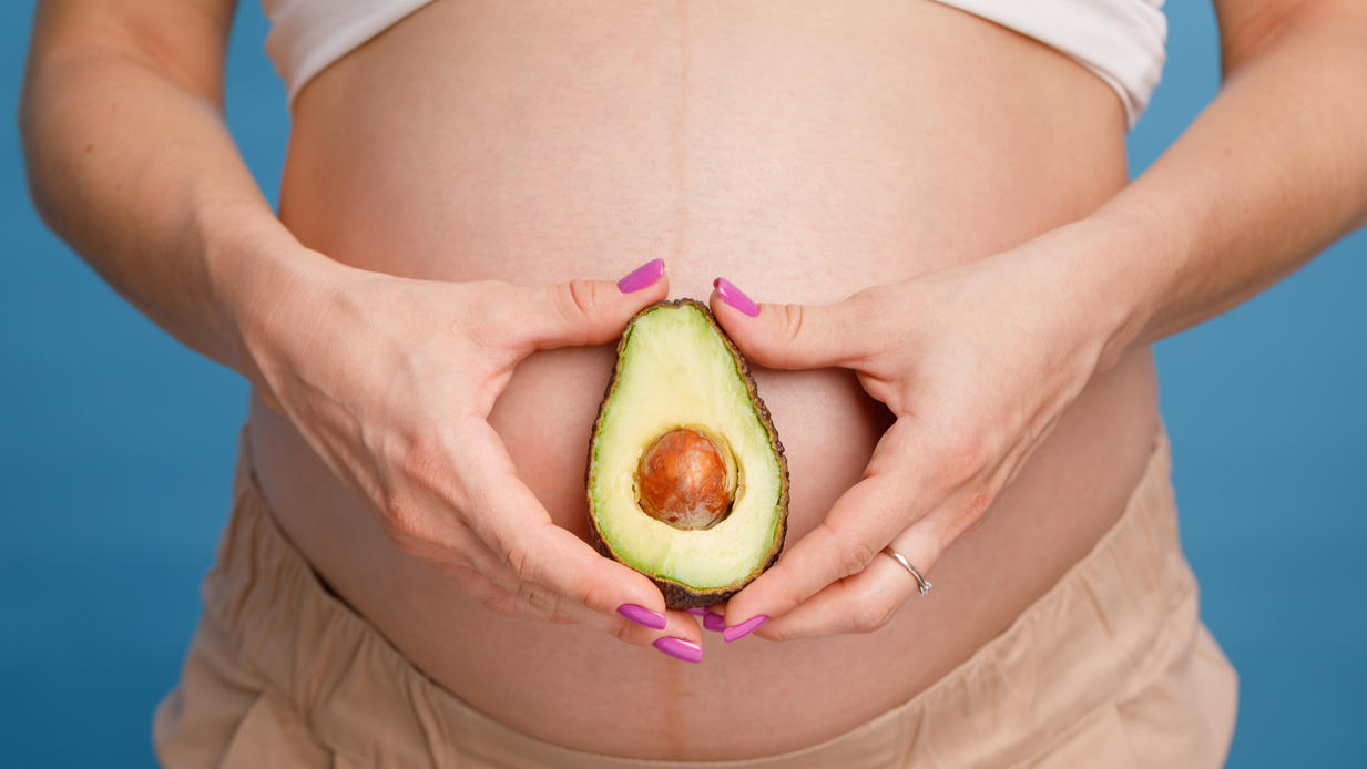 7 Lebensmittel, die wie die Organe aussehen, für die sie gut sind! Schwangere Frau hält Avocado vor ihren Bauch.