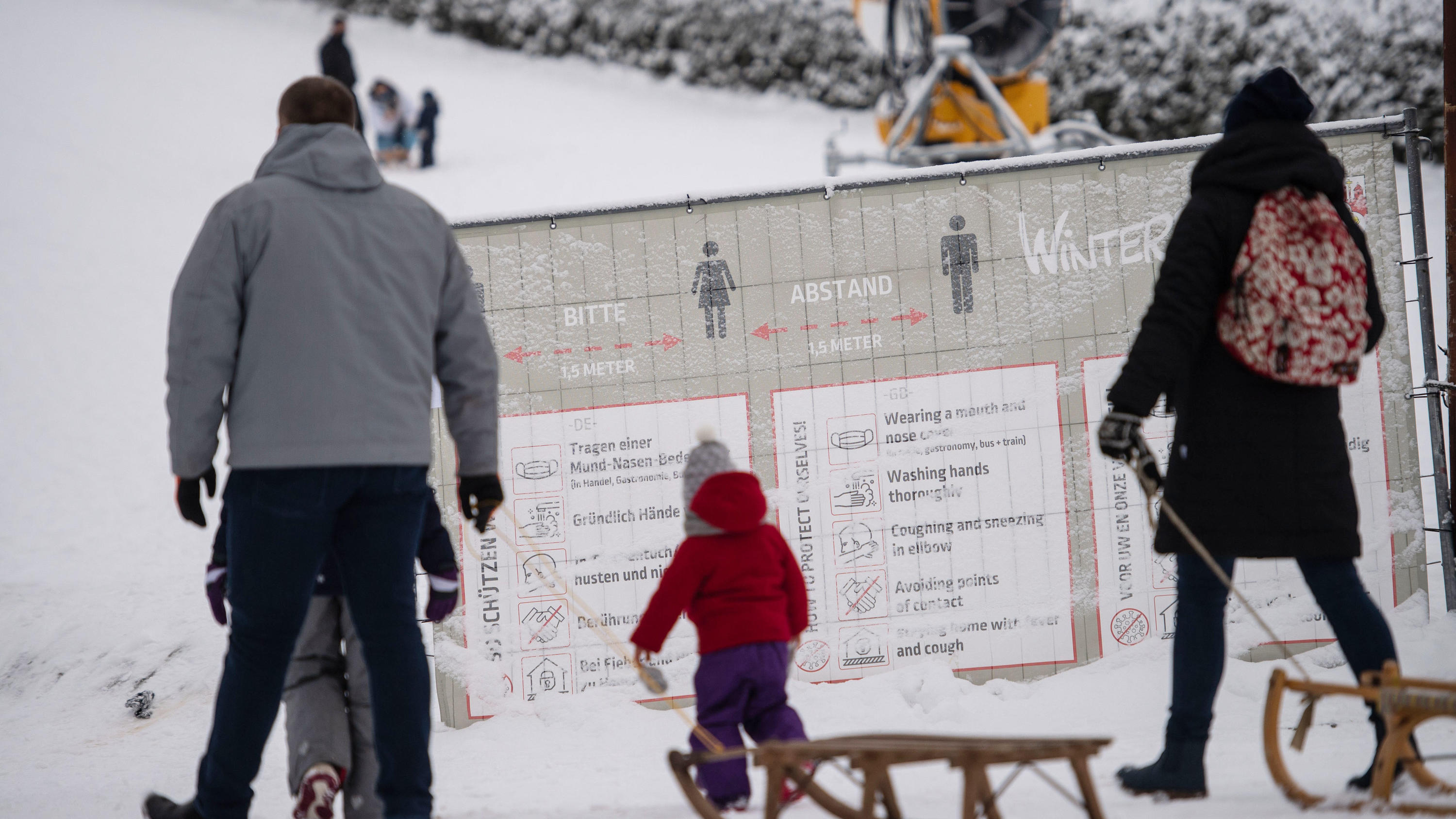 Ansturm auf Deutschlands Skigebiete - Polizei appelliert: 01.01.2021, Nordrhein-Westfalen, Winterberg: Ausflüger mit Schlitten gehen an einer geschlossenen Skipiste an einem Banner mit Corona-Verhaltensregeln vorbei. Die Stadt Winterberg bittet wegen der Corona-Pandemie auf Besuche in dem Wintersportort und