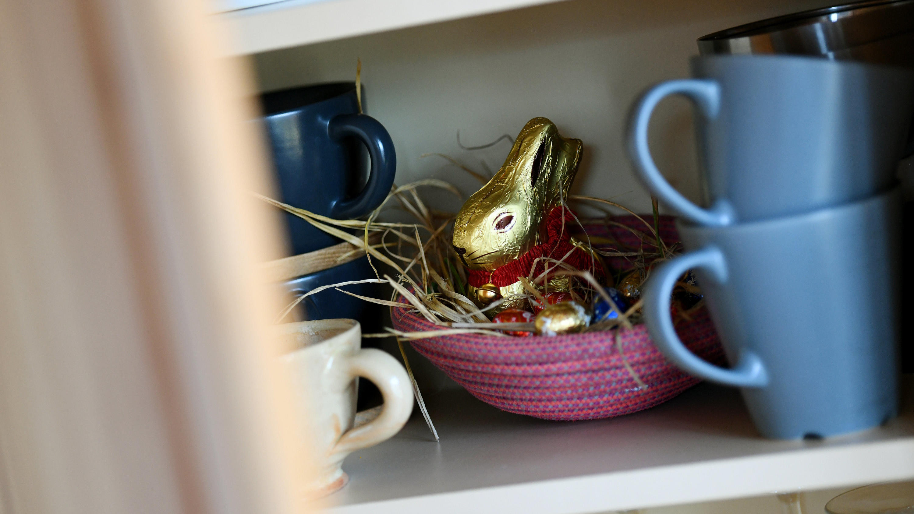 Das Wetter im März: Der erste Monat im Frühling zieht alle Register ILLUSTRATION - 10.04.2020, Bayern, Garmisch-Partenkirchen: Ein Osternest mit Schokohase und Schokoladeneiern ist in einer Wohnung zwischen Geschirr im Schrank versteckt. Zum zweiten Mal schon lässt das Coronavirus Ostern anders ablaufen als gewohnt.