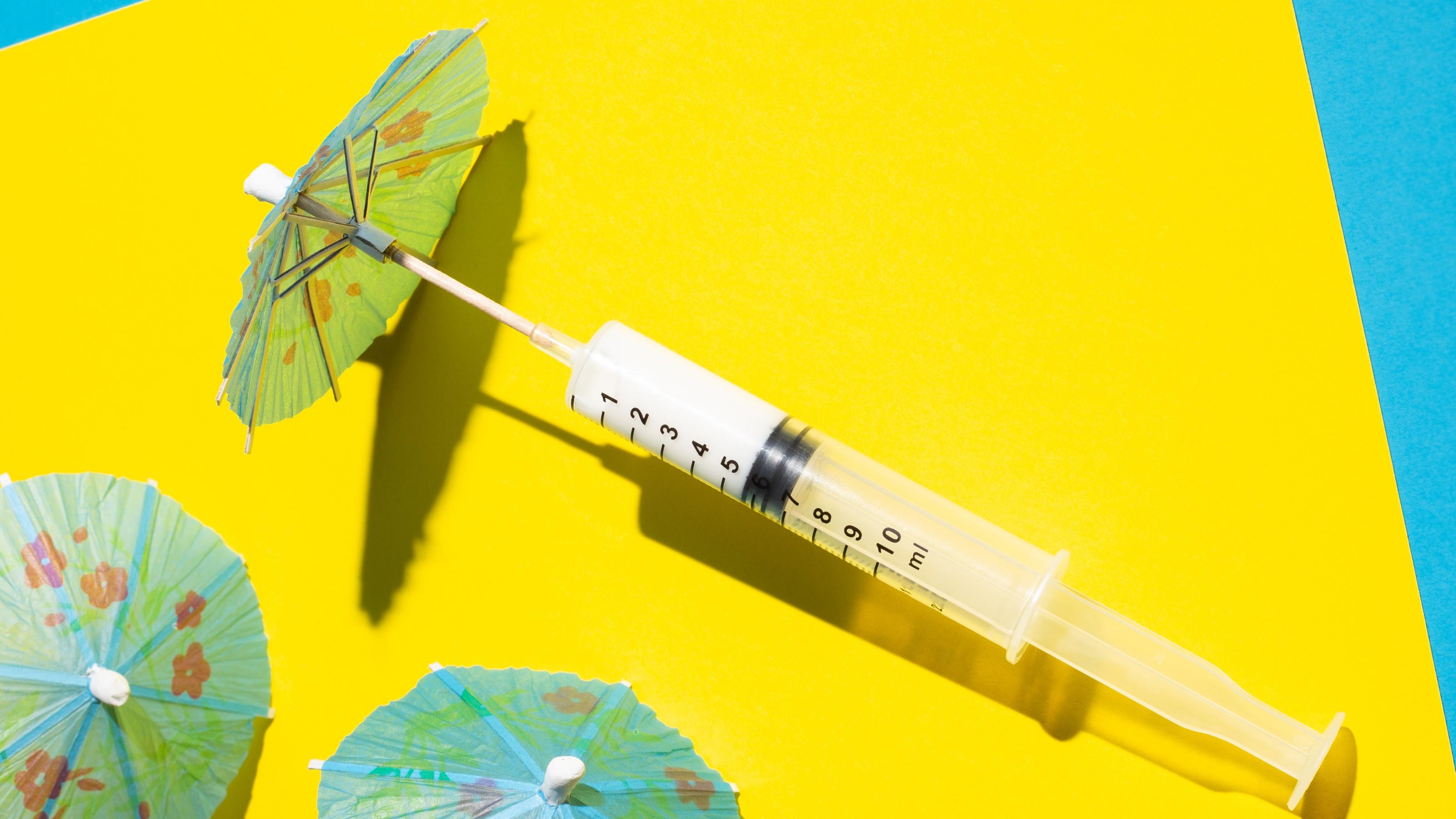 Spritz-Tour: Kuba will Urlauber mit Impfung anlocken Syringe with cocktail umbrella on the beach made of yellow and blue paper. Summer 2021 tourism and traveling concept. Minimal Coronavirus or 2019-nCoV or COVID-19 scene.