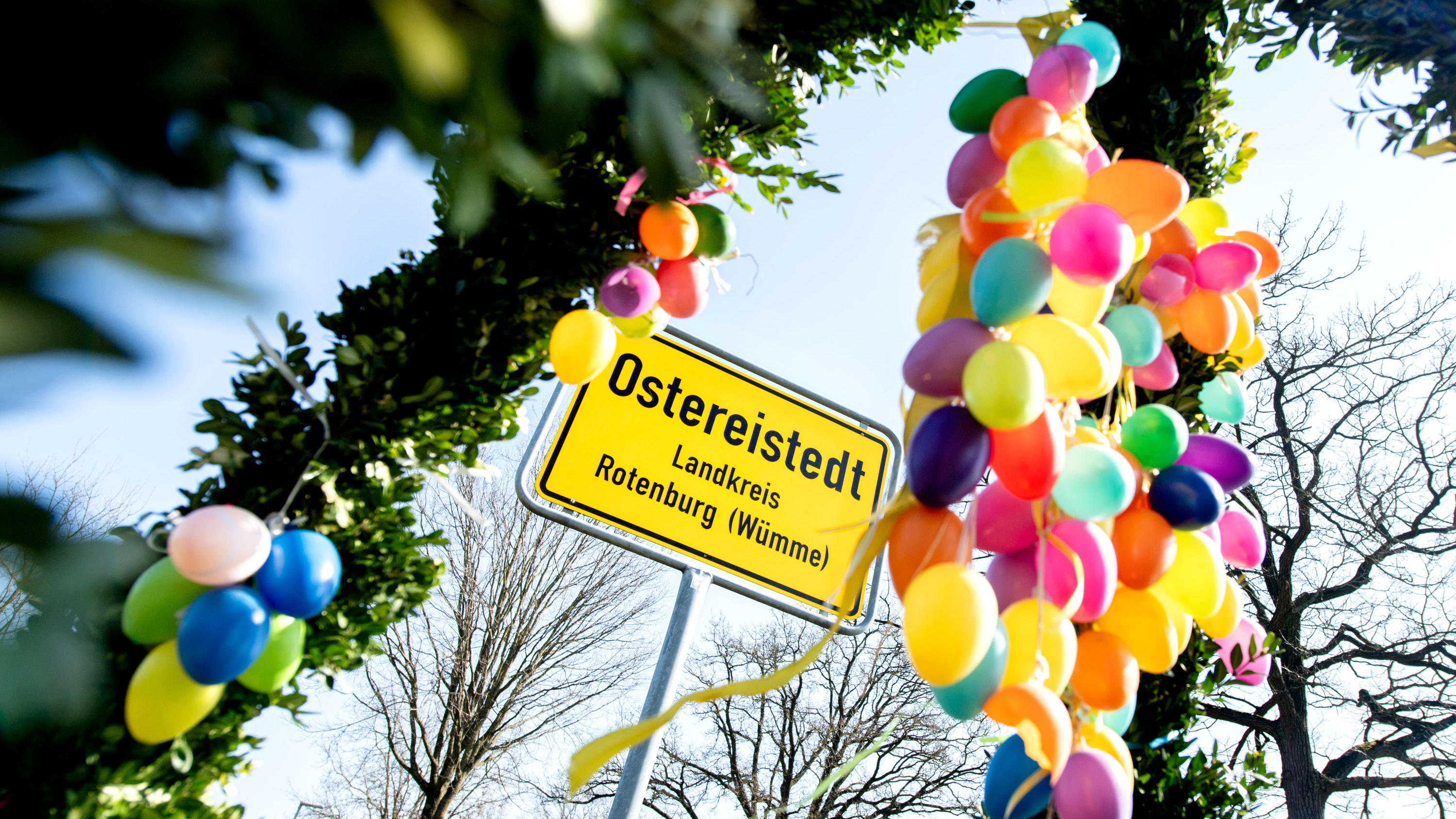 Gaaaanz viel Sonnenschein: Hier wird Ihr Osterfest schön 31.03.2021, Niedersachsen, Ostereistedt: Bunte Ostereier hängen an einem Strauch vor dem Ortsschild am Ortseingang der Gemeinde. Foto: Hauke-Christian Dittrich/dpa +++ dpa-Bildfunk +++