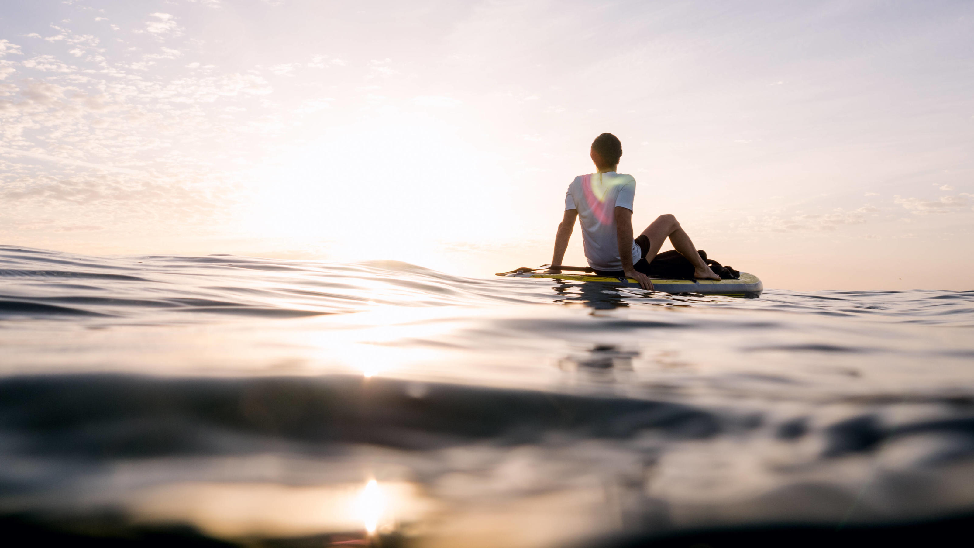 20-5-3-Regel verspricht weniger Stress im Alltag Auf dem offenen Meer entspannt ein Mann auf einem Surfboard, man sieht im Hintergrund wie die Sonne untergeht.