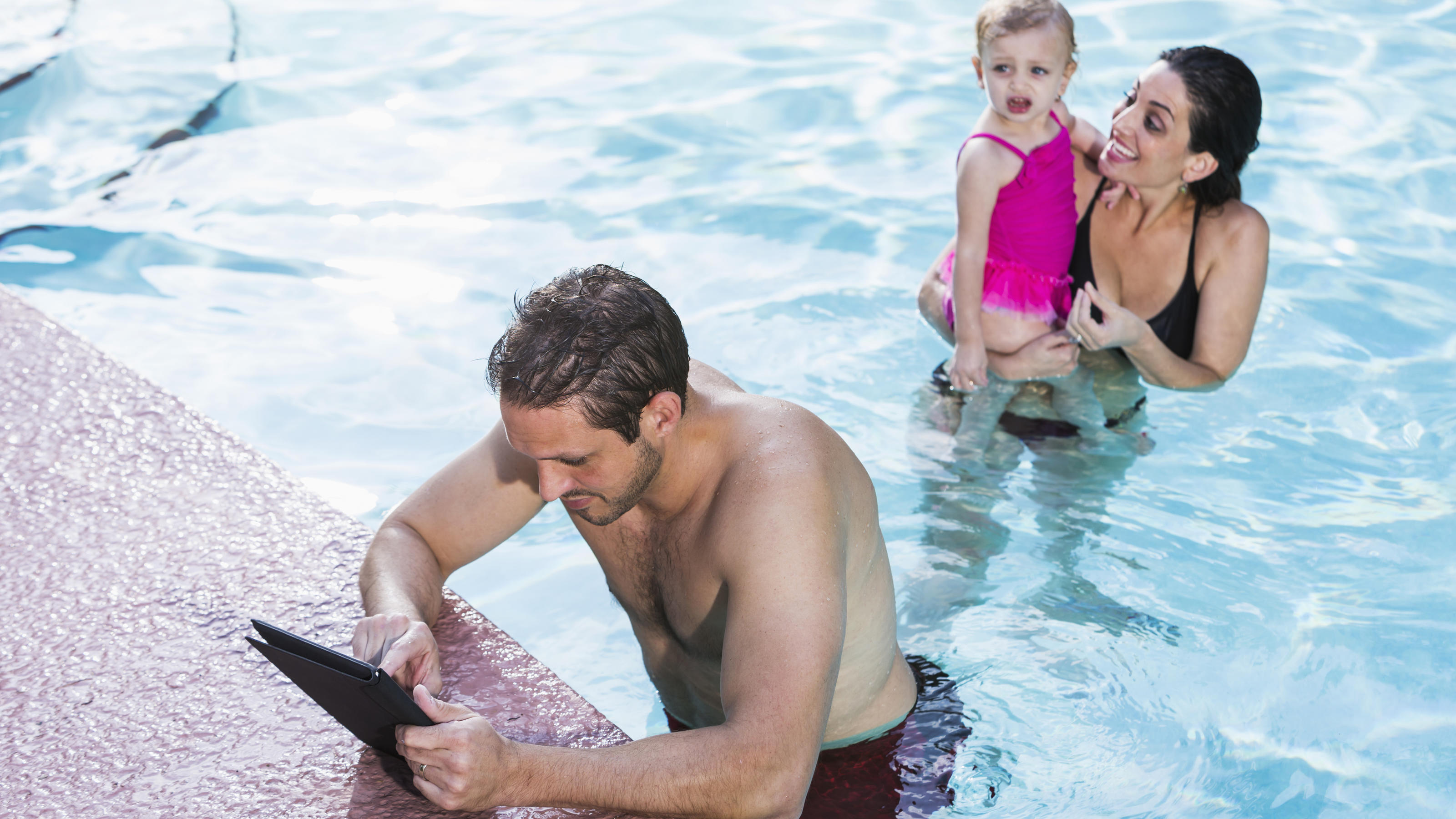 Schwimmbad-Betreiber warnt: Eltern lassen sich zu sehr vom Handy ablenken Ein Mann hängt im Schwimmbad am Beckenrand und schaut auf sein iPad, im Hintergrund sieht man eine Frau mit Kind.
