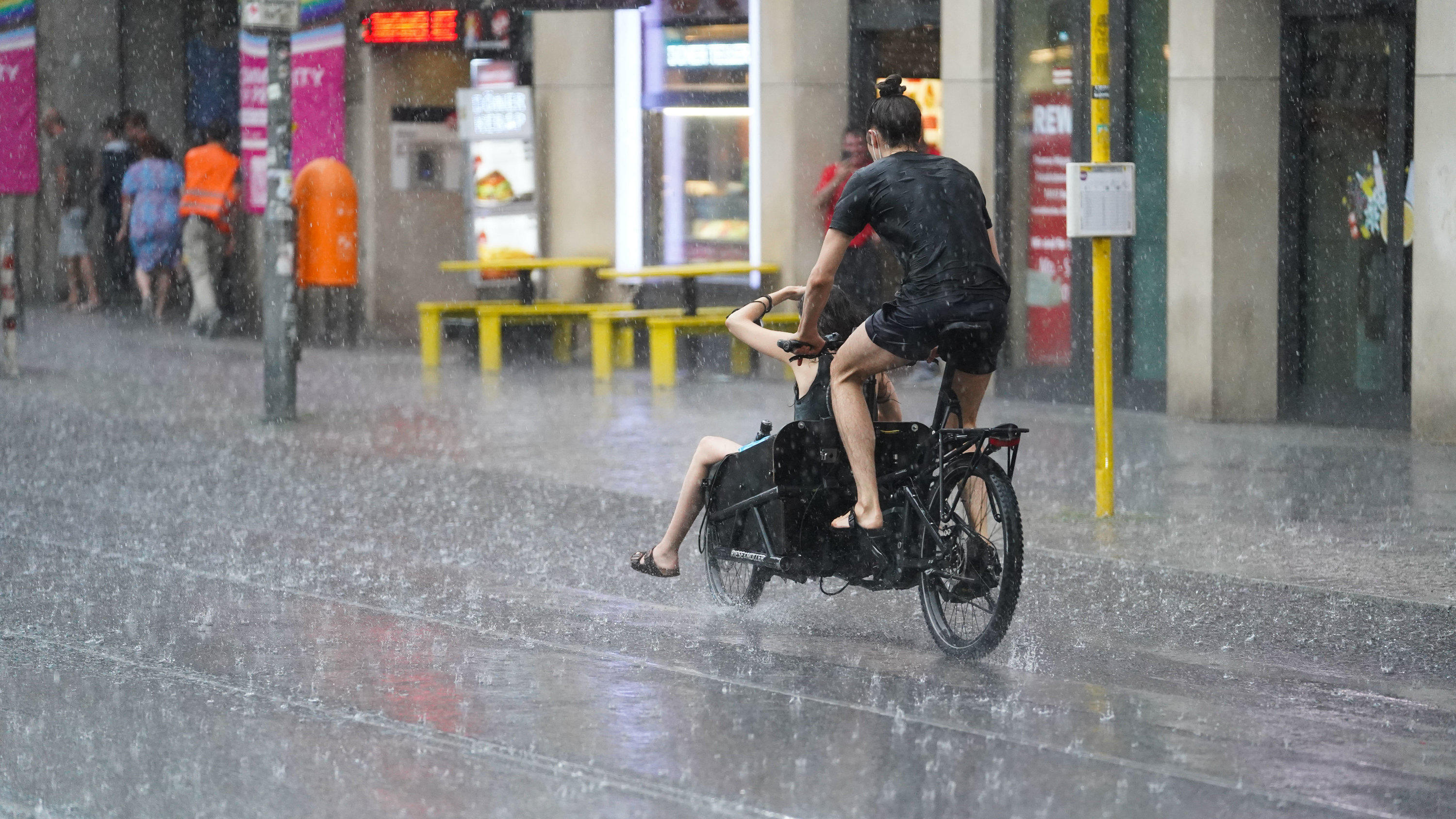 Gewitter mit Starkregen von den Alpen bis in den Osten Deutschlands 25.07.2021, Berlin: Junge Leute fahren mit einem Lastenfahrrad, während starker Regen auf die Straße prasselt.
