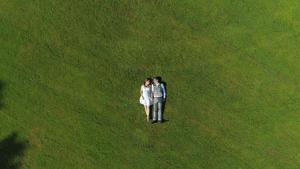 AERIAL TOP DOWN: Happy young couple laying in grass on summer day. Man and wife newlyweds relaxing in sunny park after wedding. Young man and pregnant woman lying and holding hands, expecting a baby