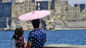 26.07.2019, Italien, Neapel: Eine Familie schützt sich mit einem Schirm vor der Sonne vor dem Castel dell'Ovo. Foto: Ciro Fusco/ANSA/AP/dpa +++ dpa-Bildfunk +++