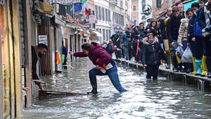15.11.2019, Italien, Venice: Eine Frau versucht, eine überflutete Straße zu überqueren, während Menschen bei Hochwasser auf einer Gerüstbrücke laufen. Nach dem schweren Hochwasser in Venedig ist die Lage weiterhin angespannt. Foto: Andrea Merola/ANSA/dpa +++ dpa-Bildfunk +++