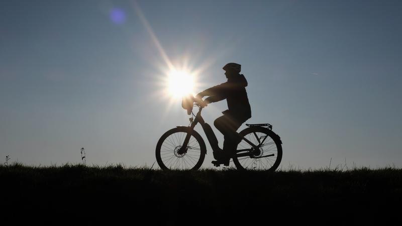 Eine Frau fährt bei untergehender Sonne mit ihrem Fahrrad über den Deich am Rhein. Foto: Martin Gerten/dpa +++ dpa-Bildfunk +++