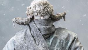 A determined frozen Caucasian male looking at the camera, all bundled up in a fur trappers hat, parka, scarf and gloves, wearing glasses and carrying a yellow snow shovel, covered in snow and frost from the driving snow of a blizzard, heads out to shovel snow.