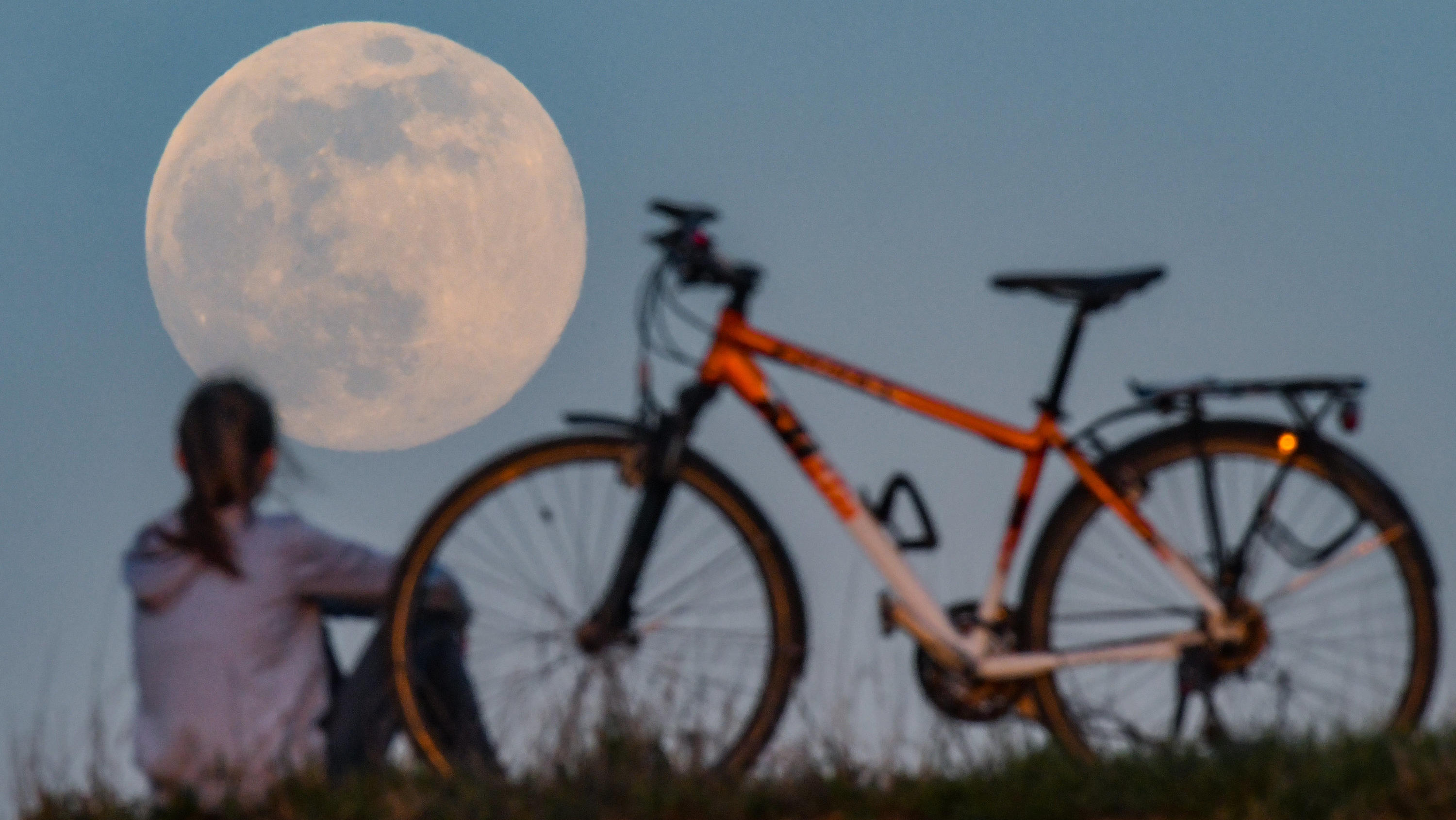 07.04.2020, Brandenburg, Sieversdorf: Der Mond geht als sogenannter Supermond auf, während auf einem Hügel ein Mädchen neben ihrem Fahrrad sitzt und das Schauspiel bewundert. Der Mond erreicht in der Nacht vom 07. auf den 08. April als Vollmond sein 