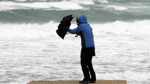 A woman in a blue raincoat holding a black umbrella struggles with the umbrella due to stormy, windy weather on a beach