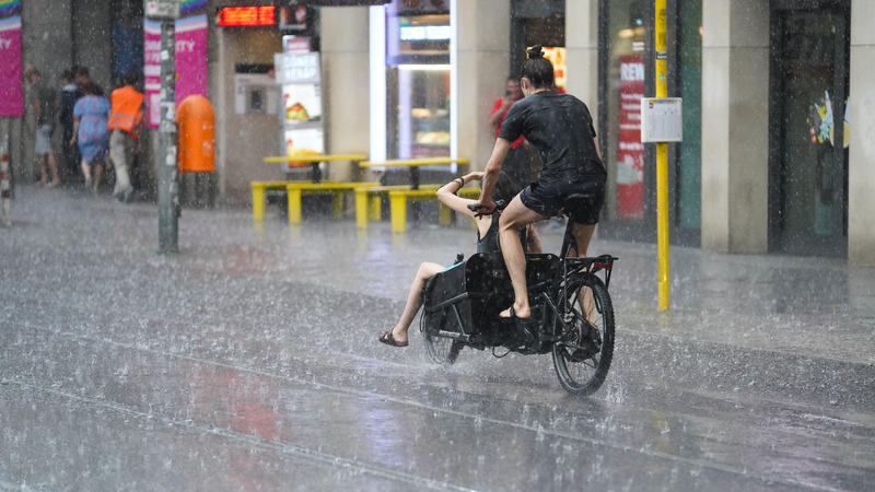 25.07.2021, Berlin: Junge Leute fahren mit einem Lastenfahrrad, während starker Regen auf die Straße prasselt.