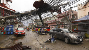 19.12.2021, Philippinen, Surigao: Autos fahren an einem durch den Taifun Rai umgestürzten Strommast in der Stadt Surigao, Surigao del Norte, im Süden der Philippinen, vorbei, da die Stromversorgung unterbrochen ist. Die Zahl der Todesopfer des stärksten Taifuns, der die Philippinen in diesem Jahr heimgesucht hat, steigt weiter an, und der Gouverneur einer Inselprovinz, die vom Taifun Rai besonders hart getroffen wurde, sagte, dass es möglicherweise noch größere Verwüstungen gibt, über die noch nicht berichtet wurde. Foto: Jilson Tiu/AP/dpa +++ dpa-Bildfunk +++