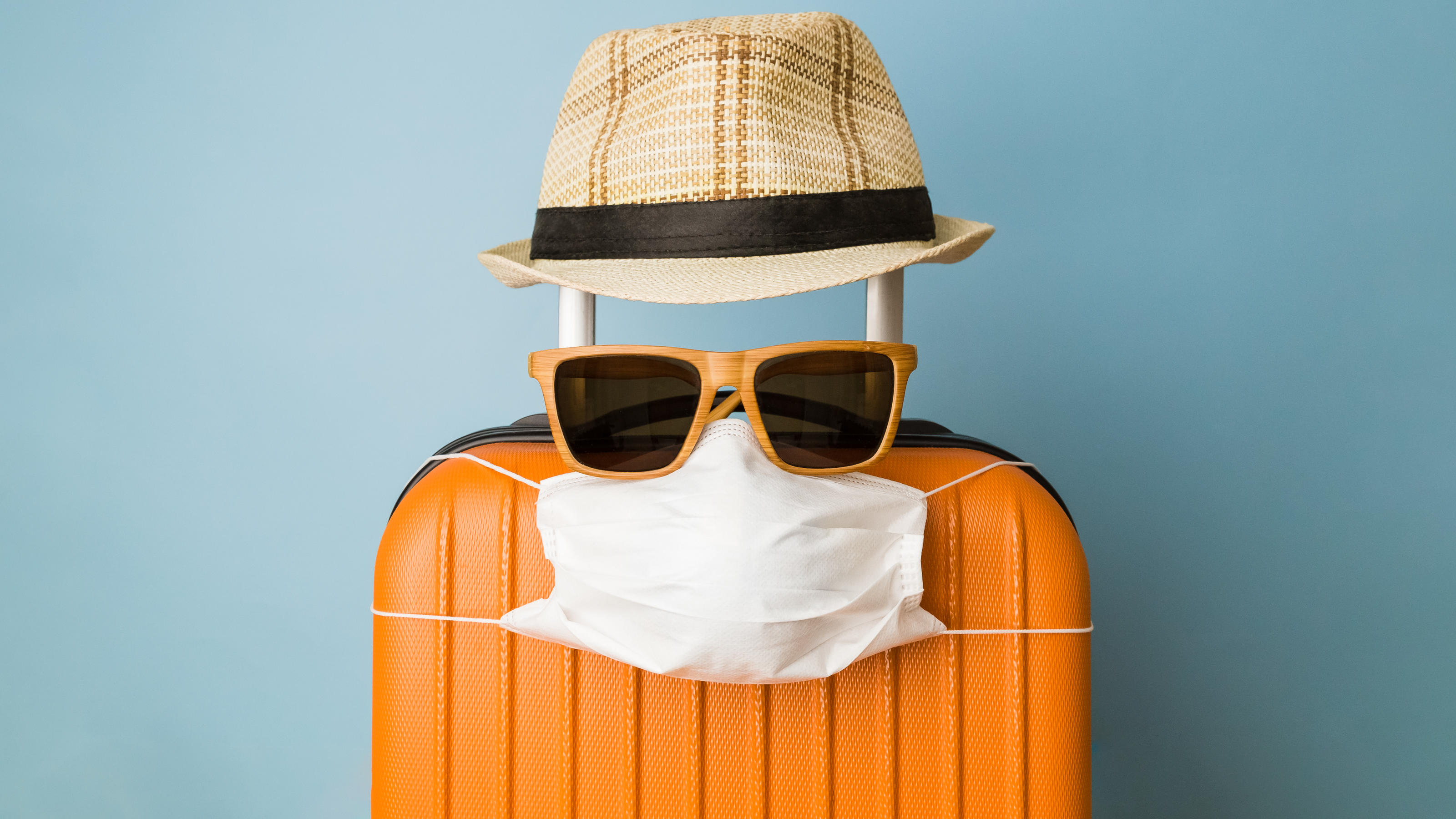 Ein Koffer "trägt" eine Sonnenbrille und eine Corona-Maske.