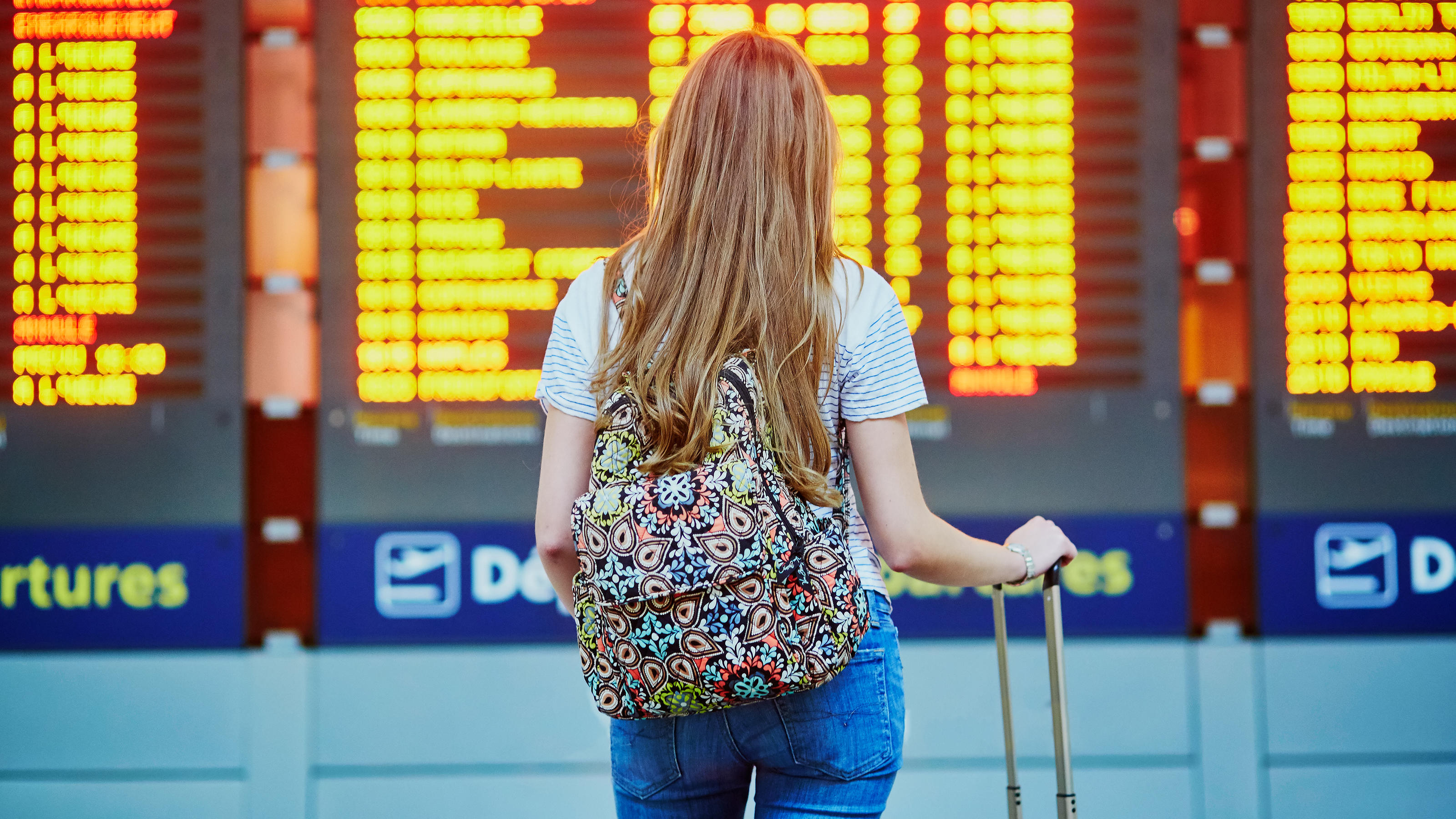 Beautiful young tourist girl with backpack and carry on luggage in international airport, near flight information board