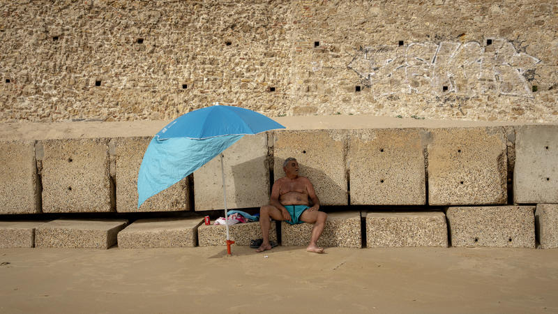 13.06.2022, Spanien, Cadiz: Ein Mann sitzt auf einem Wellenbrecher und schützt sich vor der Sonne unter seinem Sonnenschirm an einem Strand. Große Teile Spaniens werden in dieser Woche von einer für Juni ungewohnt heftigen Hitzewelle heimgesucht. Die