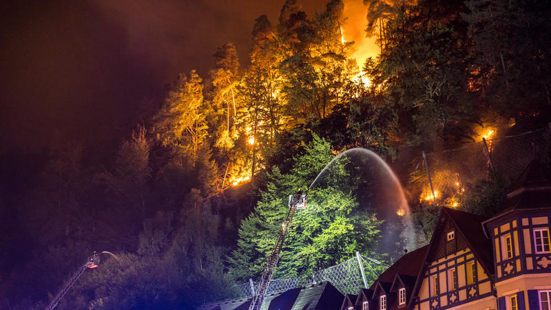 26.07.2022, Tschechien, Hrensko: Flammen lodern direkt hinter Häusern während ein Feuerwehrmann veruscht mit seinem Schlauch zu löschen. Ein Waldbrand im tschechischen Nationalpark Böhmische Schweiz nahe der Grenze zu Sachsen hat weiter die Einsatzkr