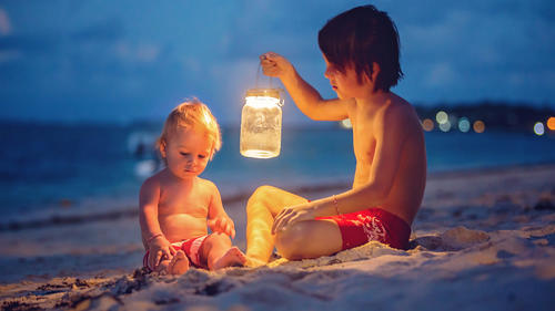 Little toddler boy, sitting on the beach after sunset on a moonlight with lantern, enjoying the quietness of the beach on tropical island