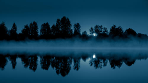 Night mystical scenery. Full moon through the tree branches, rising over the foggy river and its reflection in the still water.