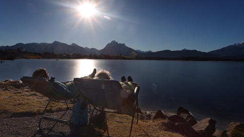 ARCHIV - 15.02.2023, Bayern, Füssen: Zwei Frauen genießen am Ufer des am Alpenrand gelegenen Hopfensees auf Liegestühlen den Sonnenschein. Die Menschen in Deutschland sind trotz wirtschaftlicher Sorgen einer Umfrage zufolge in Reiselaune. (zu dpa: «Reiseanalyse deutet auf starke Urlaubsnachfrage trotz Inflation hin») Foto: Karl-Josef Hildenbrand/dpa +++ dpa-Bildfunk +++