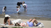 04.06.2023, Nordrhein-Westfalen, Rodenlirchen: Zwei Frauen liegen am Strand des Rhein und genießen das warme Wetter mit Sonnenschein, andere Menschen kühlen sich im kühlen Nass des Flusses ab. Die Meteorologen sagen die ganze nächste Woche Sommerwetter an. Foto: Roberto Pfeil/dpa +++ dpa-Bildfunk +++