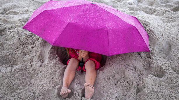03.08.2023, Mecklenburg-Vorpommern, Boltenhagen: Bei kräftigem Regen sitzt ein Ferienkind unter seinem Schirm am Strand. Kühl und regnerisch zeigt sich das Wetter derzeit in Norddeutschland. Foto: Jens Büttner/dpa +++ dpa-Bildfunk +++