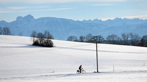 29.11.2023, Bayern, Ruderatshofen: Ein Radfahrer fährt im Sonnenschein vor dem Panorama der Alpen durch die verschneite Landschaft. Foto: Karl-Josef Hildenbrand/dpa +++ dpa-Bildfunk +++