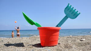 Symbolbild zum Thema Strandurlaub mit Kindern: In einem roten Plastikeimerchen stecken eine kleine grüne Schaufel und ein blauer Rechen, aufgenommen an einem Strand bei Genua (Provinz Ligurien) am 20.07.2009. Foto: Jochen Lübke +++(c) dpa - Report+++