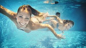 Group of mid 20's people taking underwater selfies in a swimming pool. There are two girls and a guy in foreground, keeping airtight, smiling and looking at camera.