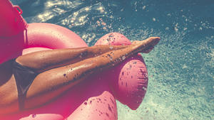 Woman floating on a pink inflatable in swimming pool. Tight crop showing only her legs and feet. She is tanned in turquoise water with water splashing onto her.